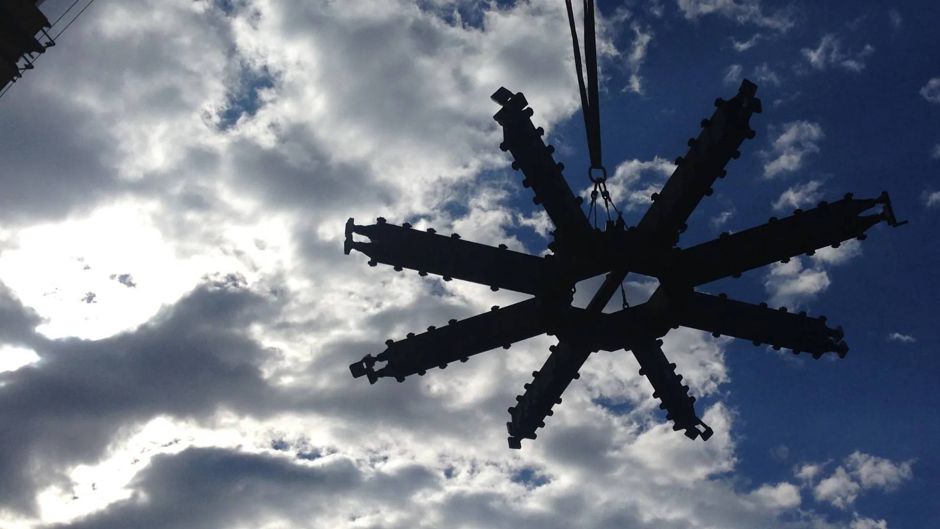 A multi-arm portable machining fixture suspended from a crane against a cloudy sky.